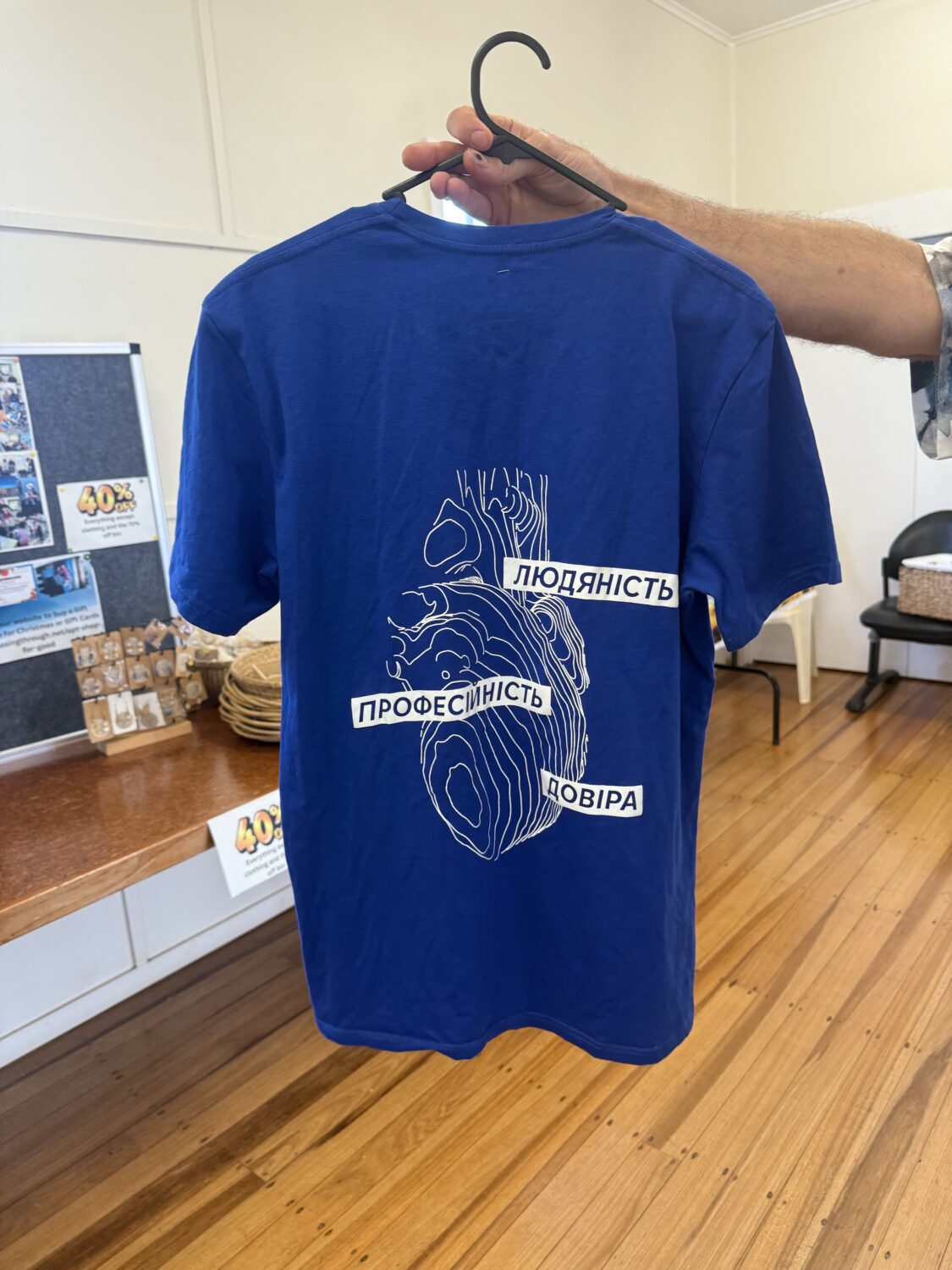 Blue T-shirt on a hanger featuring a white abstract heart design and Ukrainian words Людяність, Професійність, Довіра.