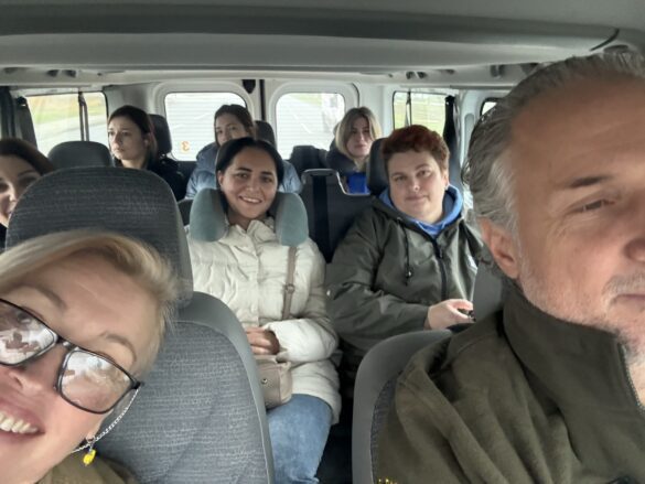 IMG_6547-585×439-2 Smiling group of eight adults seated in a van, taking a casual selfie from the front passenger area.