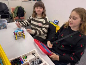 Two girls sit at a table building a small colorful LEGO robot; one holds a pink tablet while the other watches nearby.