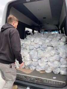 Man in a dark hoodie stands beside an open van packed with dozens of white plastic bags inside and spilling onto the floor at the ramp edge