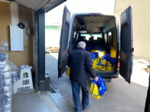 Man in a dark jacket stands at the back of an open van, carrying blue-and-yellow bags toward a pile of bags inside the rear cargo area.