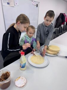 Three children at a table making crepes, brushing batter onto a round crepe on a plate with toppings nearby on the counter.