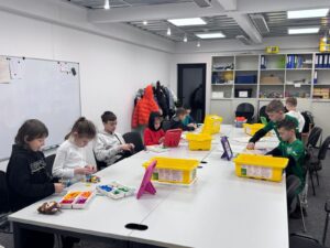 Group of children at a long table building with colorful plastic bricks in a bright classroom, with yellow storage bins nearby.