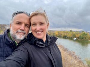 Smiling couple taking a selfie on a hillside overlooking a river with autumn trees and cloudy skies behind them.