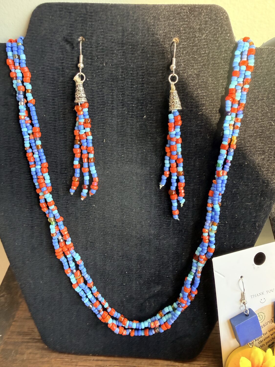 IMG_3075 Multi-strand beaded necklace in red, blue, and turquoise with silver bell charms; matching beaded earrings on a black display bust.