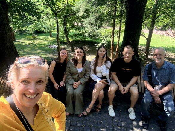 Smiling woman in yellow takes a selfie with a group of five people sitting on a bench in a sunlit park with trees behind them.