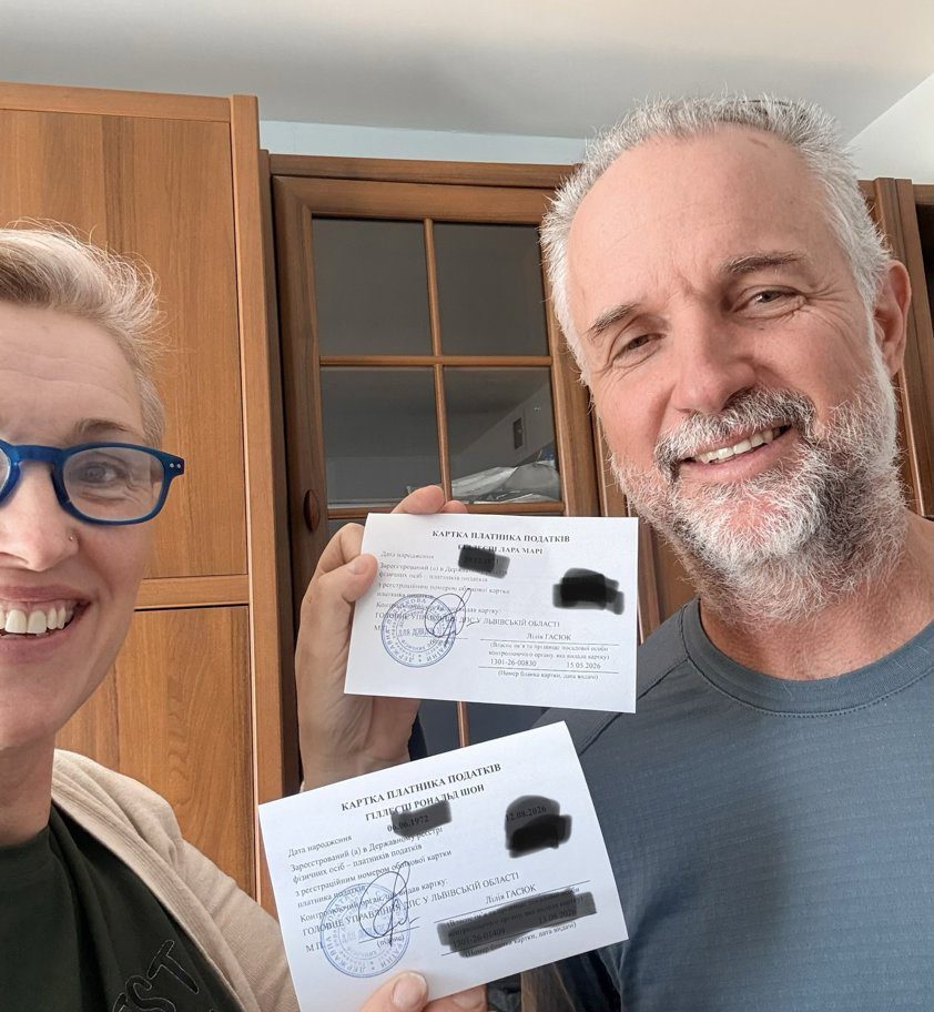 Two smiling adults indoors, each holding stamped Ukrainian tax documents/card.