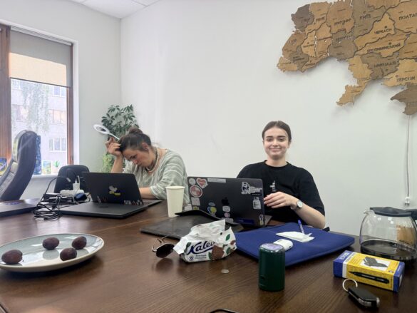 Two women sit at a wooden conference table with laptops; one rests her head on her hand, the other smiles at the camera with a sticker-covered laptop nearby.