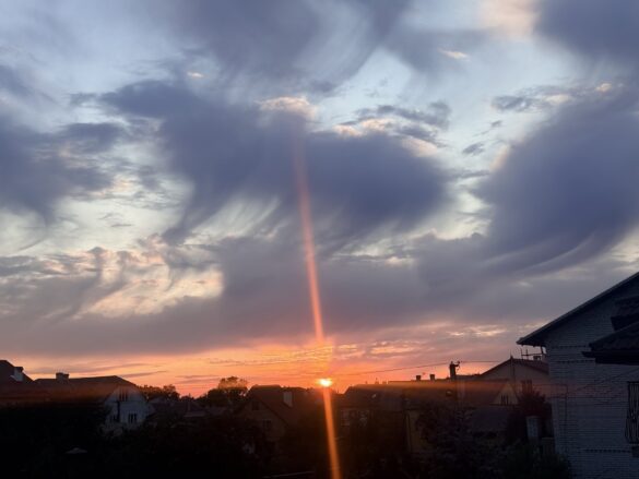 Sunset over a suburban neighborhood with orange clouds and a vertical sun pillar beam rising from the horizon behind dark silhouetted houses