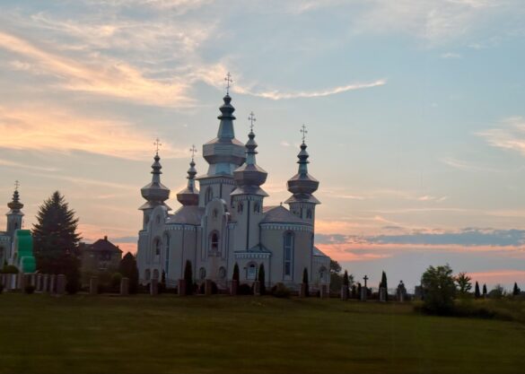 White Orthodox church with multiple onion domes and crosses at sunset, set in a grassy cemetery.