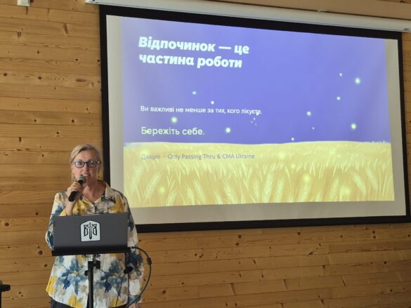 Woman with glasses speaks into a microphone at a lectern, in front of a projector screen showing a Ukrainian slide: 'Відпочинок — це частина роботи' and 'Бережіть себе', with a yellow wheat field illustration.