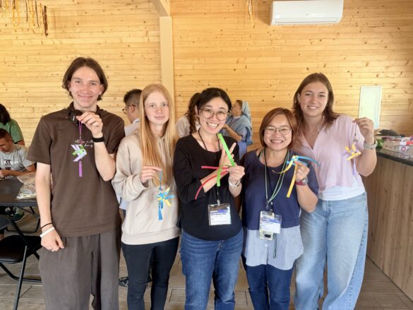 Five smiling young adults stand together in a wood-paneled room, each holding colorful paper ribbons from a craft activity.