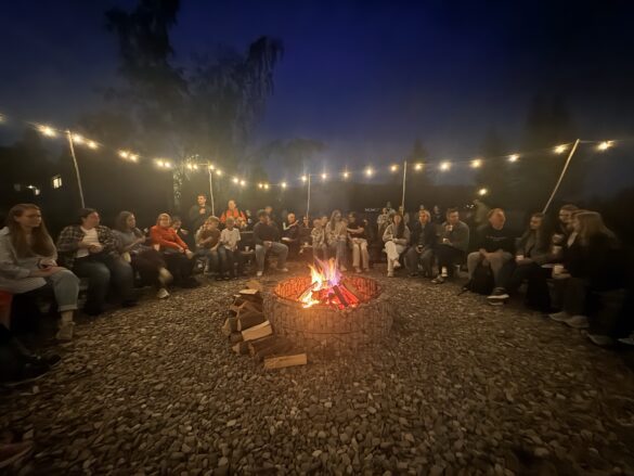 Group of people seated in a circle around a lit campfire on a gravel surface at night, with string lights overhead.