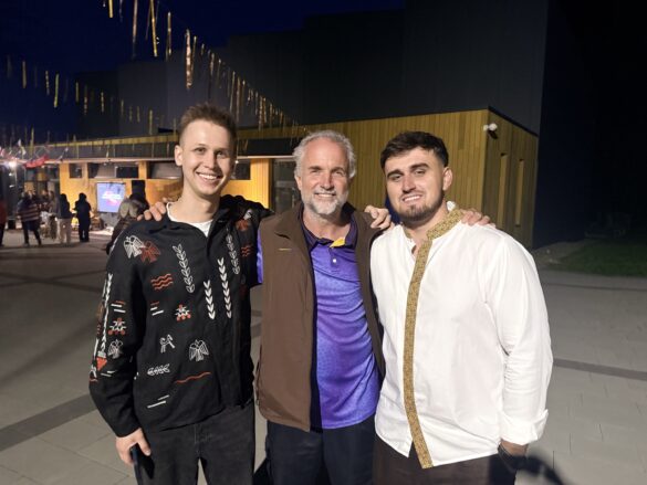 Three men posing together at night, smiling, with string lights and a wooden building in the background.