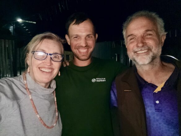 Three friends pose for a selfie at night, smiling at the camera: woman with glasses on the left, man in green shirt center, man in purple shirt and brown vest on the right.